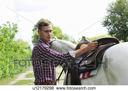 Stock Photo - Portrait of young man saddling horse. Fotosearch