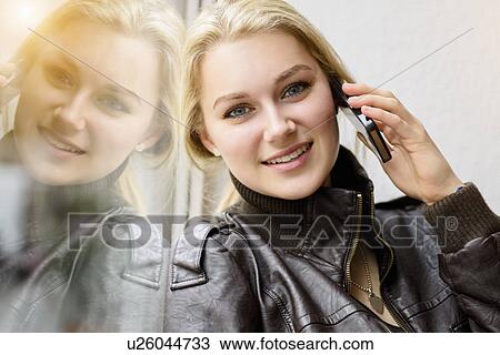 Stock Image - Portrait of young woman chatting on smartphone by window. Fotosearch