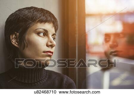 Stock Image - Portrait of young woman gazing out of window at the street. Fotosearch