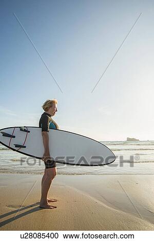 Stock Image - Senior woman standing on beach with surfboard, Camaret-sur-mer, Brittany, France. Fotosearch