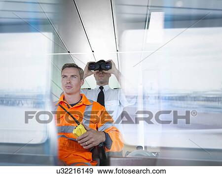 Stock Photo - Ship worker and captain using binoculars on ship's bridge with reflections on window. Fotosearch