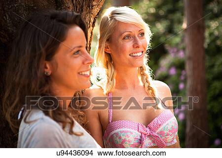 Picture - Two female friends leaning against tree in forest. Fotosearch
