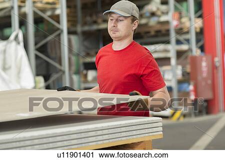 Stock Image - Worker lifting boards in hardware store warehouse. Fotosearch
