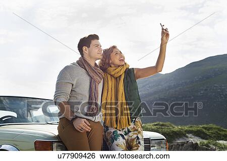 Stock Photograph - Young couple leaning against car taking selfie on smartphone, Cape Town, Western Cape, South Africa. Fotosearch