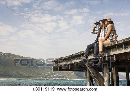 Young couple sitting on edge of old pier looking through binoculars, Cape Town, Western Cape, South Africa View Large Photo Image Stock Photo - Young couple sitting on edge of old pier looking through binoculars, Cape Town, Western Cape, South Africa. Fotosearch