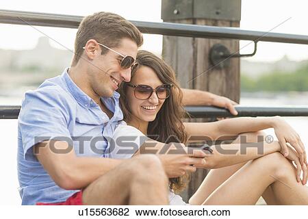 Stock Image - Young couple wearing sunglasses, man using cell phone. Fotosearch
