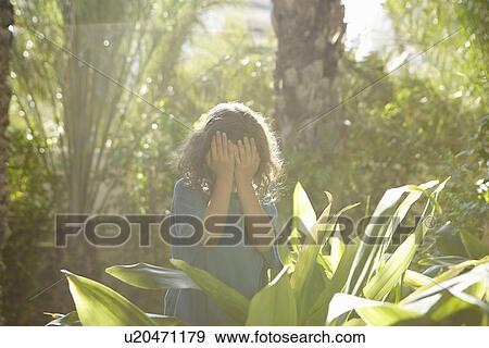 Stock Photo - Young girl, hands covering eyes, standing among trees and foliage. Fotosearch