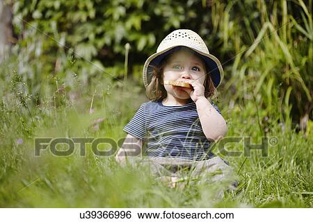 Stock Photograph - Young girl sitting in grass eating snack. Fotosearch