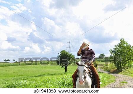 Stock Photo - Young man adjusting cowboy hat riding horse on rural road. Fotosearch
