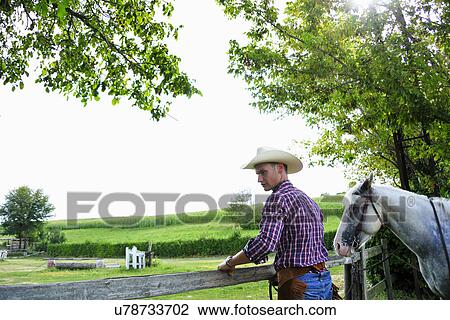 Stock Image - Young man in cowboy gear with horse checking fence. Fotosearch