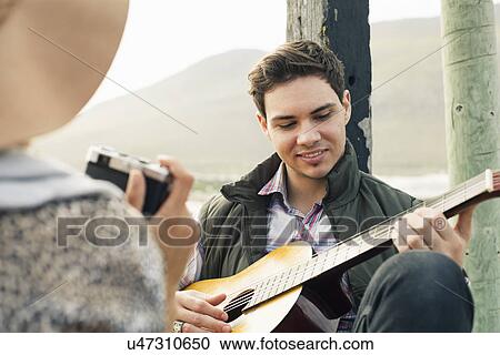 Stock Image - Young man playing acoustic guitar for girlfriend on coastal pier, Cape Town, Western Cape, South Africa. Fotosearch
