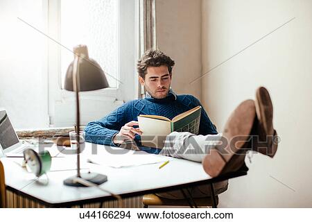 Picture - Young man reading book with feet up on desk. Fotosearch