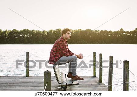 Stock Photo - Young man sitting on bench on jetty looking across water. Fotosearch
