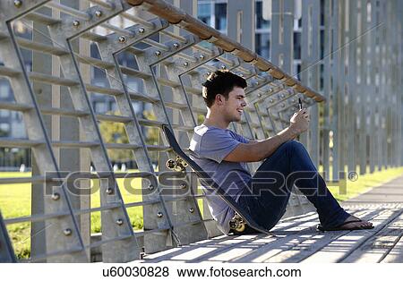 Young man sitting, using mobile phone, skateboard beside him View Large Photo Image Stock Photo - Young man sitting, using mobile phone, skateboard beside him. Fotosearch