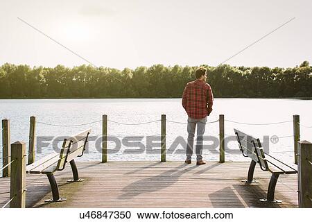 Stock Image - Young man standing on jetty looking across water. Fotosearch