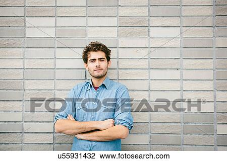 Stock Image - Young man wearing denim shirt by brick wall, portrait. Fotosearch