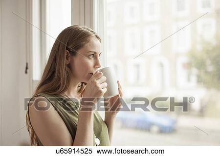 Stock Photography - Young woman drinking coffee and gazing through sitting room window. Fotosearch