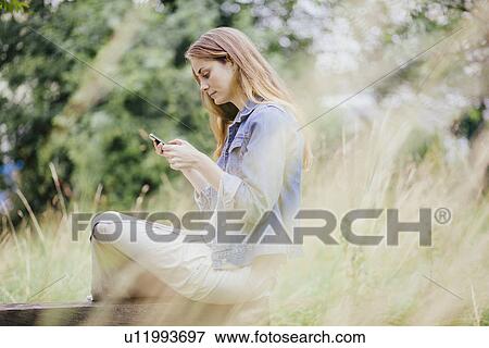 Young woman reading texts on smartphone on park bench View Large Photo Image Stock Photo - Young woman reading texts on smartphone on park bench. Fotosearch