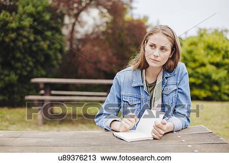 Stock Image - Young woman sitting at picnic bench in park writing in notebook. Fotosearch