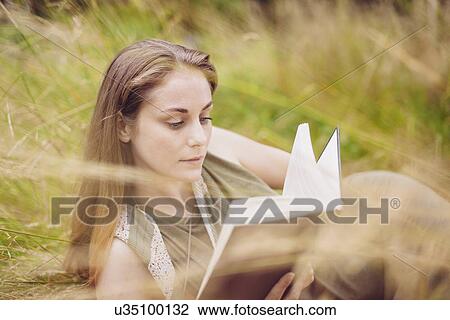 Stock Image - Young woman sitting in long grass reading book. Fotosearch