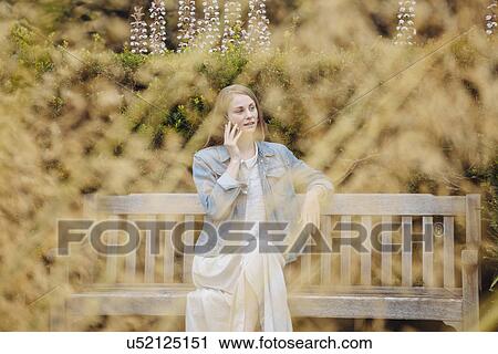 Stock Image - Young woman sitting on park bench chatting on smartphone in park. Fotosearch