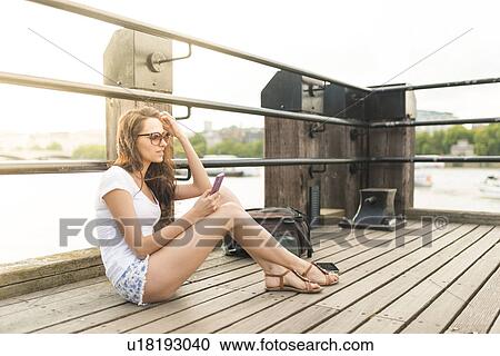 Stock Image - Young woman sitting on pier using cell phone. Fotosearch