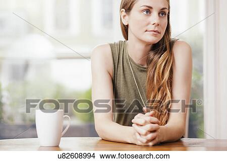 Picture - Young woman sitting with hands clasped at dining room table. Fotosearch