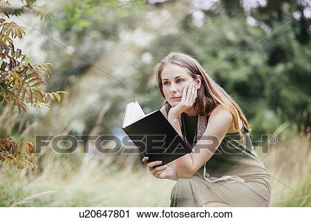 Stock Image - Young woman with chin on hand reading book in field. Fotosearch