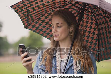 Young woman with umbrella texting on smartphone in park View Large Photo Image Stock Image - Young woman with umbrella texting on smartphone in park. Fotosearch