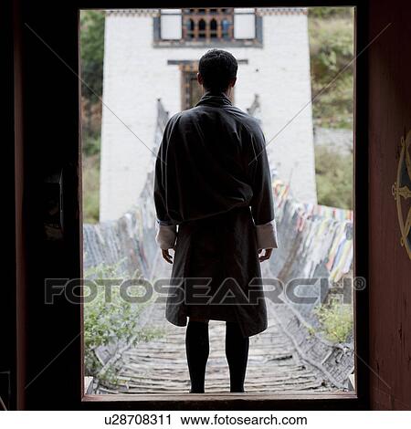 Stock Image - Bhutan man standing in the forfront of the Tamchhog Lhakhang iron suspension bridge, Bhutan. Fotosearch