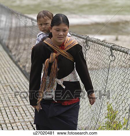 Stock Image - Woman carrying her son and crossing the bridge across Puna Tsang River, Punakha District, Bhutan. Fotosearch
