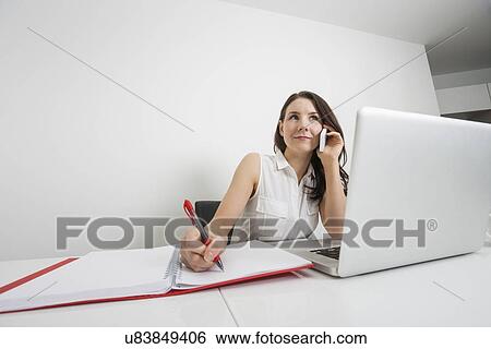 Beautiful businesswoman answering cell phone while writing at desk in office View Large Photo Image Stock Photograph - Beautiful businesswoman answering cell phone while writing at desk in office. Fotosearch