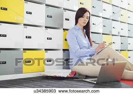 Stock Image - Beautiful businesswoman using laptop while writing in locker room at creative office. Fotosearch