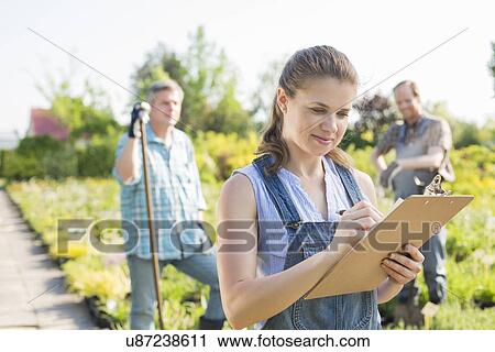 Beautiful supervisor writing on clipboard with gardeners standing in background at plant nursery View Large Photo Image Stock Image - Beautiful supervisor writing on clipboard with gardeners standing in background at plant nursery. Fotosearch