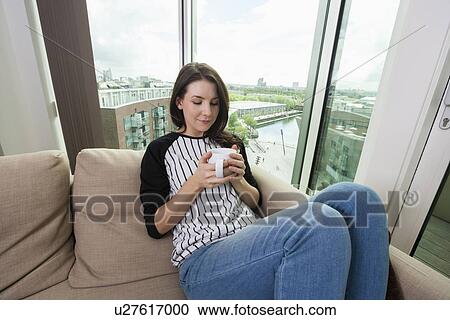 Stock Image - Beautiful woman having coffee on sofa at home. Fotosearch