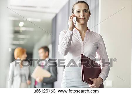 Beautiful young businesswoman on call with colleagues in background at office corridor View Large Photo Image Stock Image - Beautiful young businesswoman on call with colleagues in background at office corridor. Fotosearch