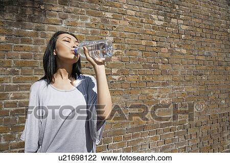 Stock Image - Beautiful young woman drinking water against brick wall. Fotosearch