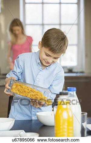 Stock Photograph - Boy pouring corn flakes in bowl with mother standing in background. Fotosearch