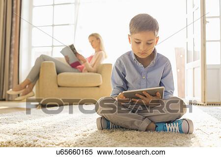 Stock Photography - Boy using digital tablet on floor with mother reading magazine in background. Fotosearch