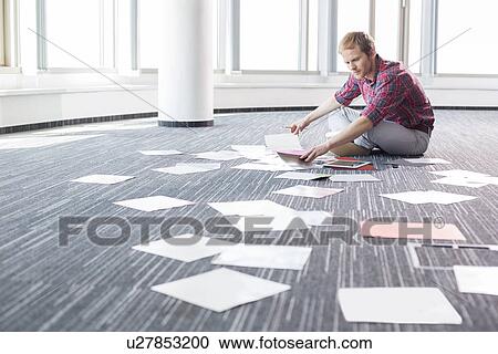 Stock Image - Businessman analyzing photographs while sitting on floor at creative office. Fotosearch