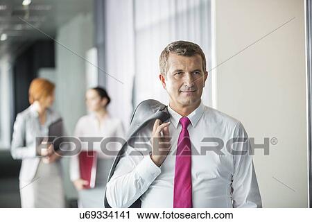 Businessman holding coat over shoulder with female colleagues in background at office View Large Photo Image Stock Image - Businessman holding coat over shoulder with female colleagues in background at office. Fotosearch
