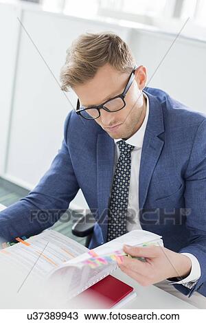 Businessman reading file at desk in creative office View Large Photo Image Stock Image - Businessman reading file at desk in creative office. Fotosearch