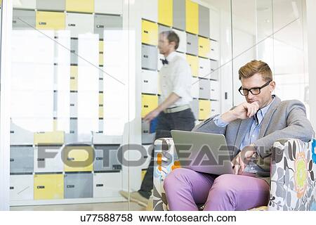 Businessman using laptop on armchair with colleague walking in background at creative office View Large Photo Image Stock Photo - Businessman using laptop on armchair with colleague walking in background at creative office. Fotosearch