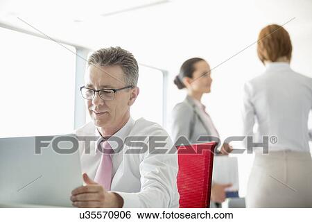 Stock Photograph - Businessman using laptop with colleagues discussing in background at office. Fotosearch