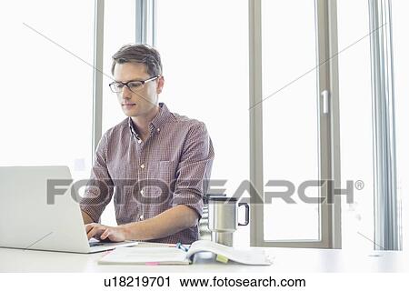 Stock Image - Businessman working on laptop at desk in creative office. Fotosearch