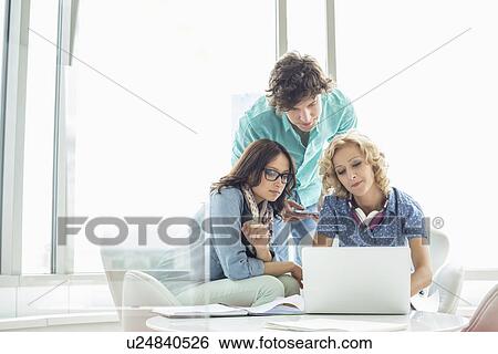 Businesspeople using laptop at table in creative office View Large Photo Image Stock Photograph - Businesspeople using laptop at table in creative office. Fotosearch