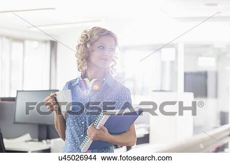 Picture - Businesswoman having coffee while holding files in creative office. Fotosearch