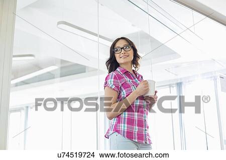 Picture - Businesswoman looking away while holding coffee mug in creative office. Fotosearch