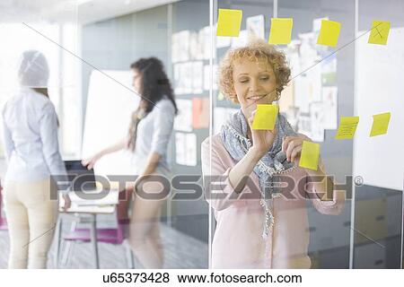 Businesswoman reading sticky notes on glass with colleagues in background View Large Photo Image Stock Photo - Businesswoman reading sticky notes on glass with colleagues in background. Fotosearch