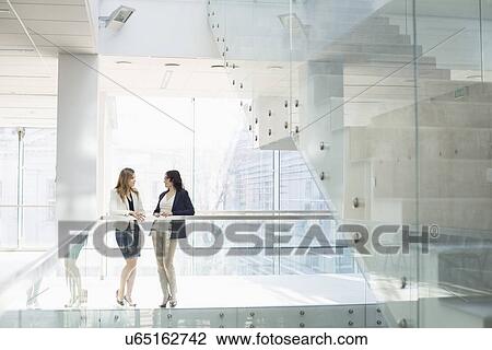 Stock Image - Businesswomen conversing against railing in office. Fotosearch